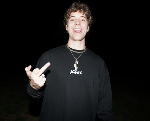 Person wearing a black KILOS sweatshirt with a logo, making a hand gesture